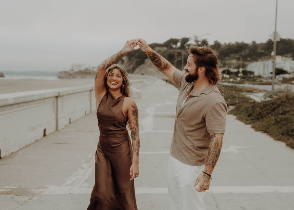 A couple dances playfully on a coastal path during sunset, with the woman wearing brown and the man in beige.