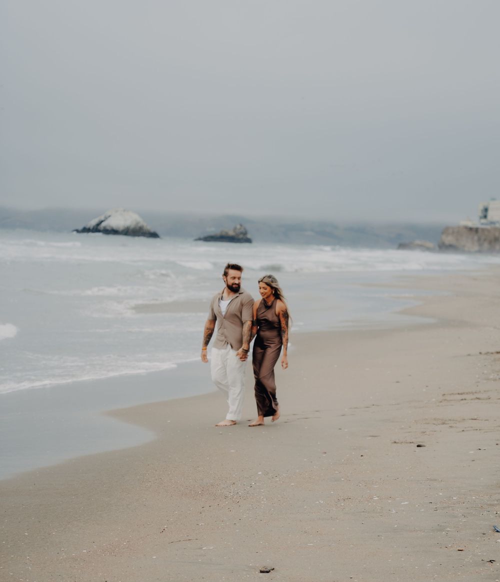 Couple taking a romantic stroll along a misty beach with waves crashing in the background.