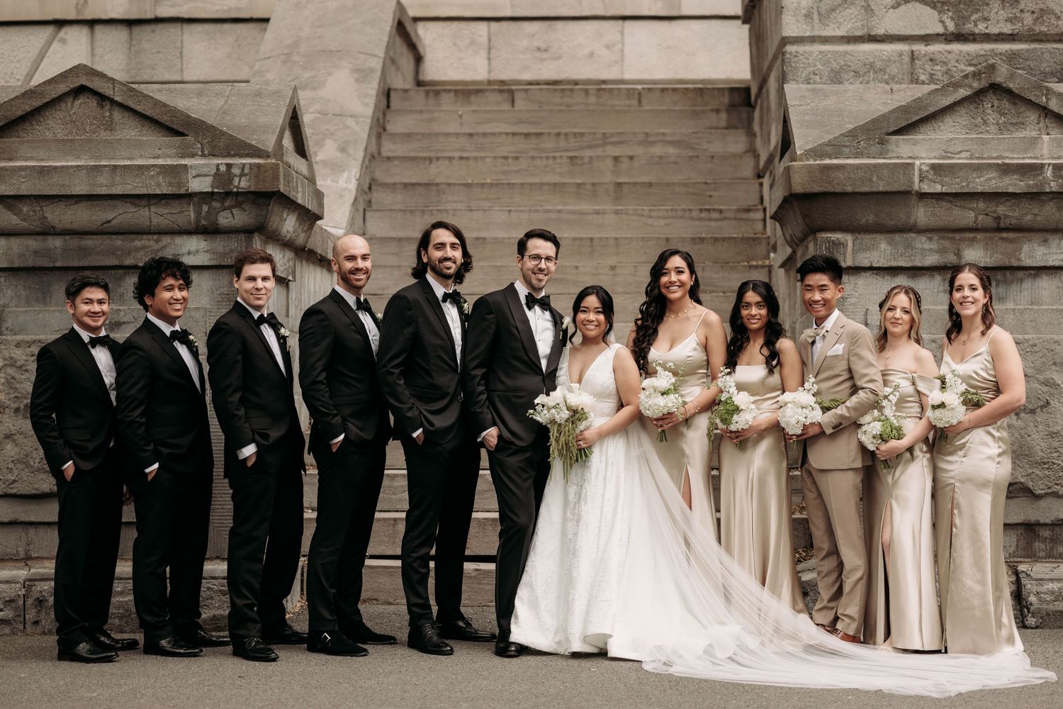 Wedding party portrait in Old Montreal. Stone steps, bride in white gown with groomsmen in black tuxedos and bridesmaids in champagne gowns.