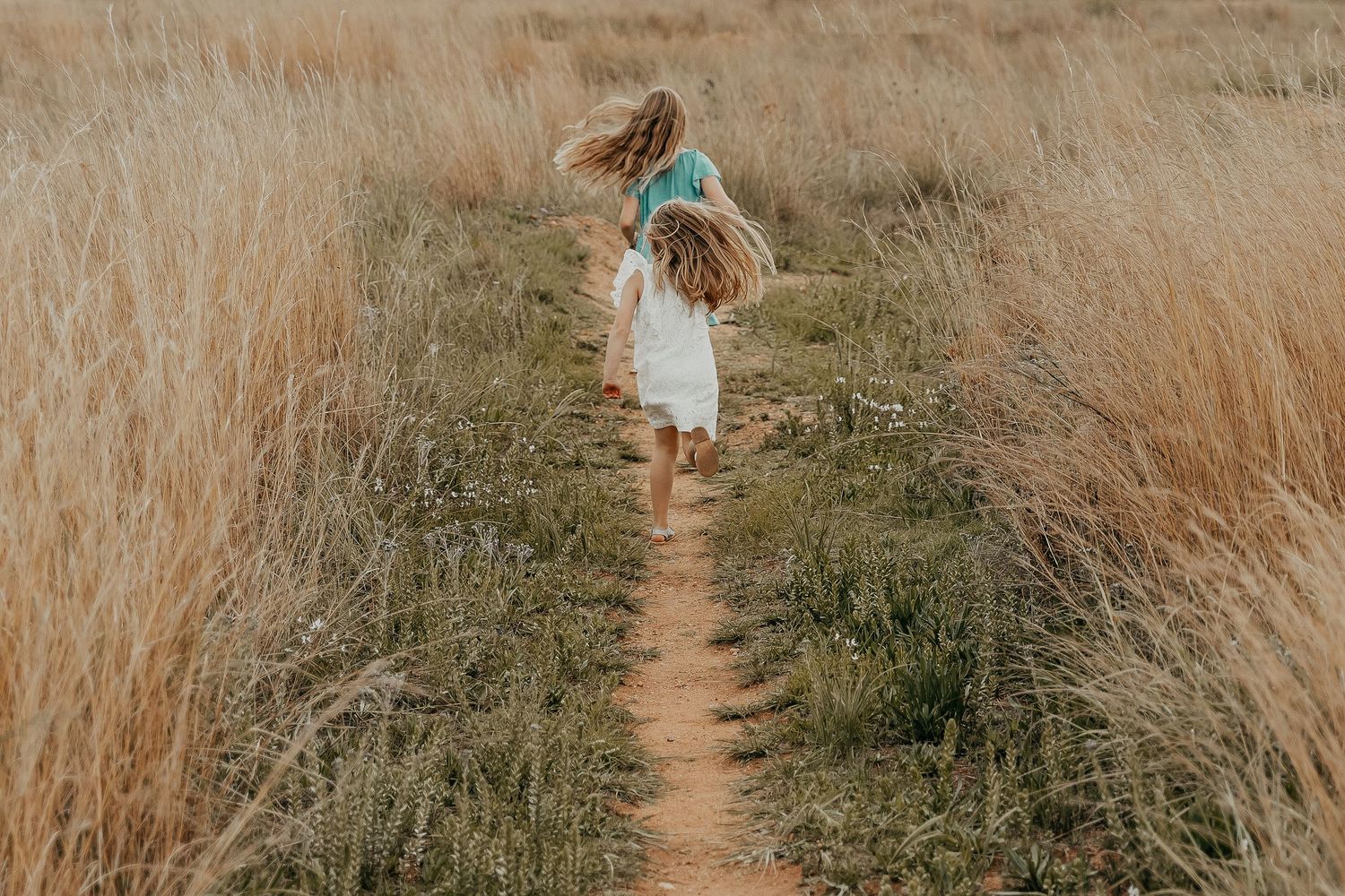 A person in a white dress walks along a winding dirt path through tall golden grass at sunset.