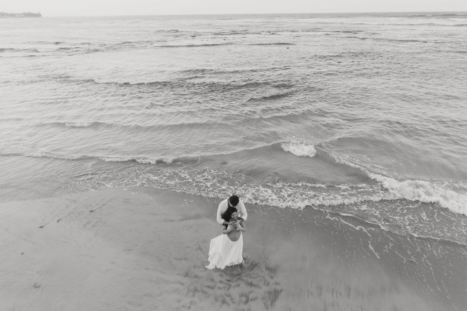 A lone figure in flowing white garments stands where gentle waves meet sandy shore.