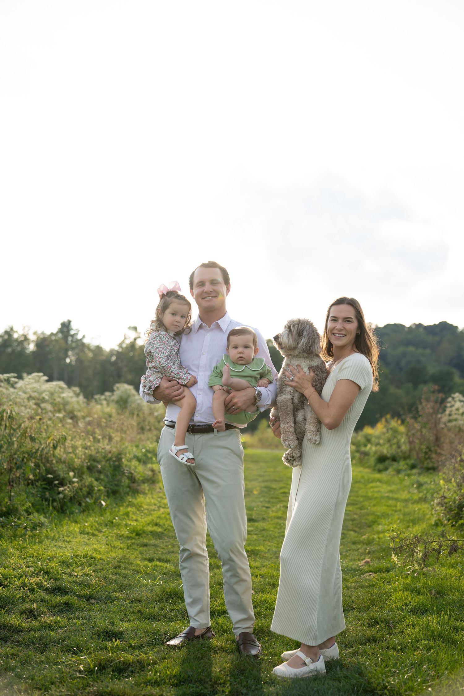 A smiling family of two adults holding two young children and a dog poses outdoors in a green meadow during golden hour.
