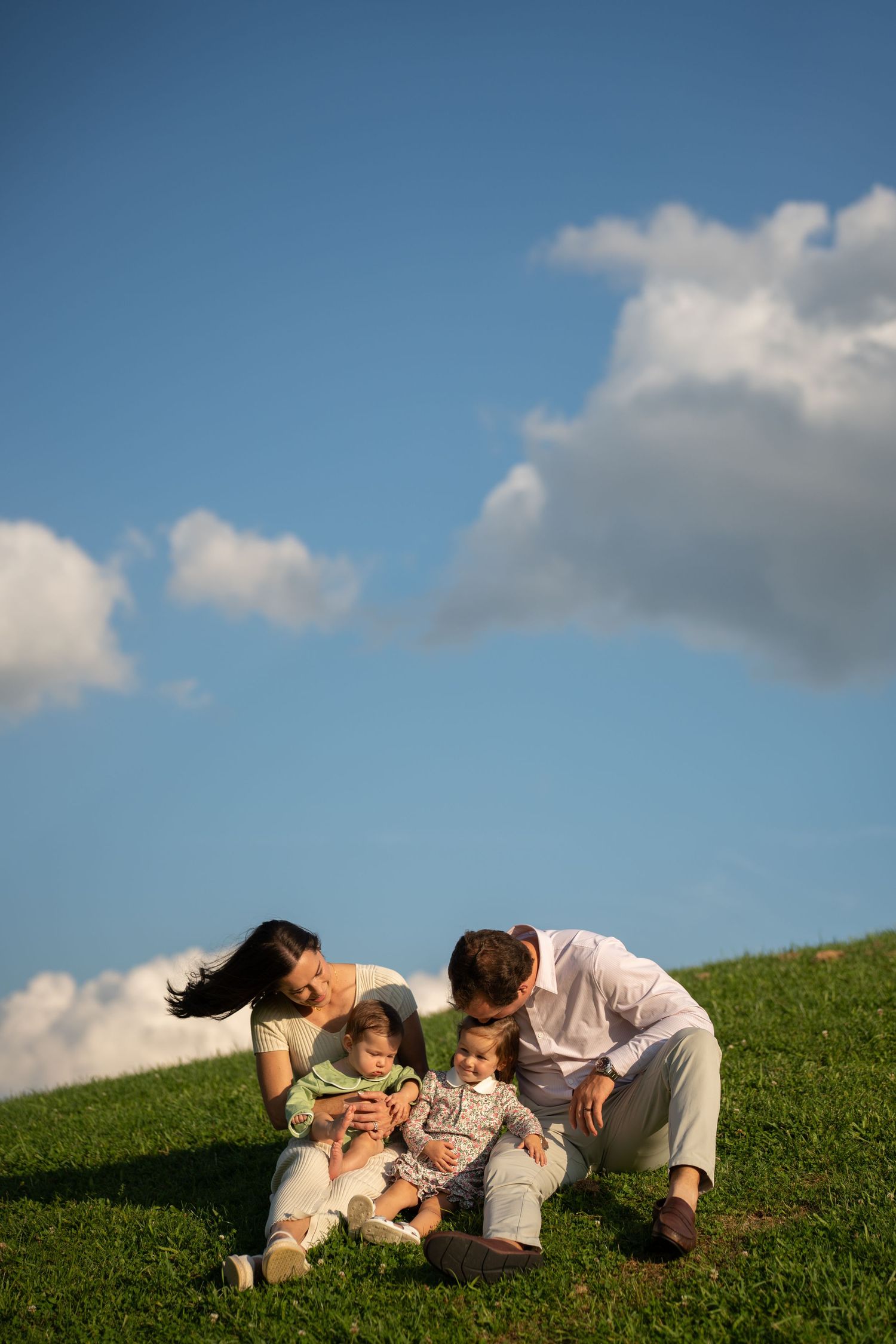 A happy family with two young children sits together on a green grassy hill under a bright blue sky with fluffy clouds.