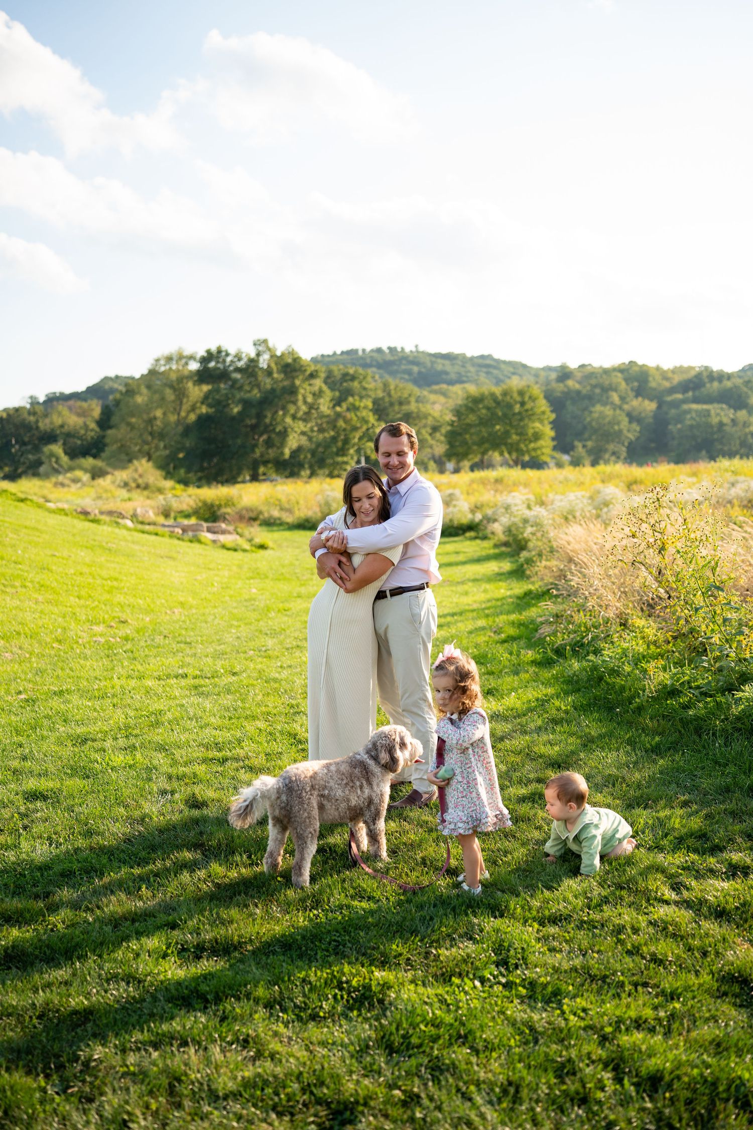 Family of four with a fluffy dog posing on a green meadow path, hugging and playing outdoors on a sunny day.