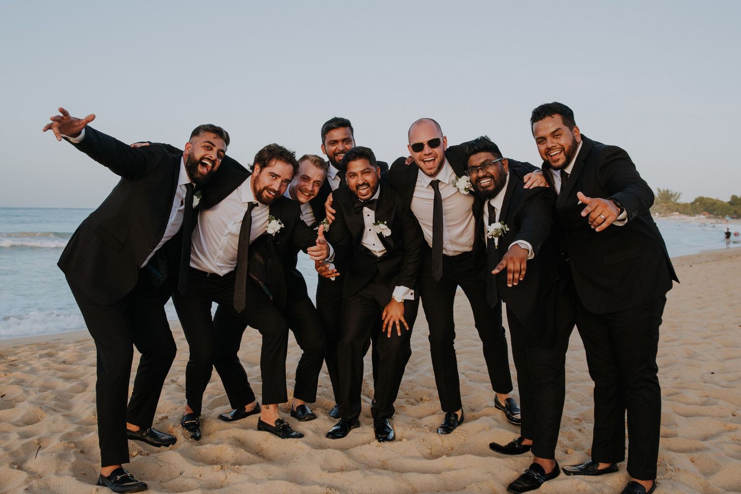 Groomsmen pose playfully on beach in black suits during sunset.