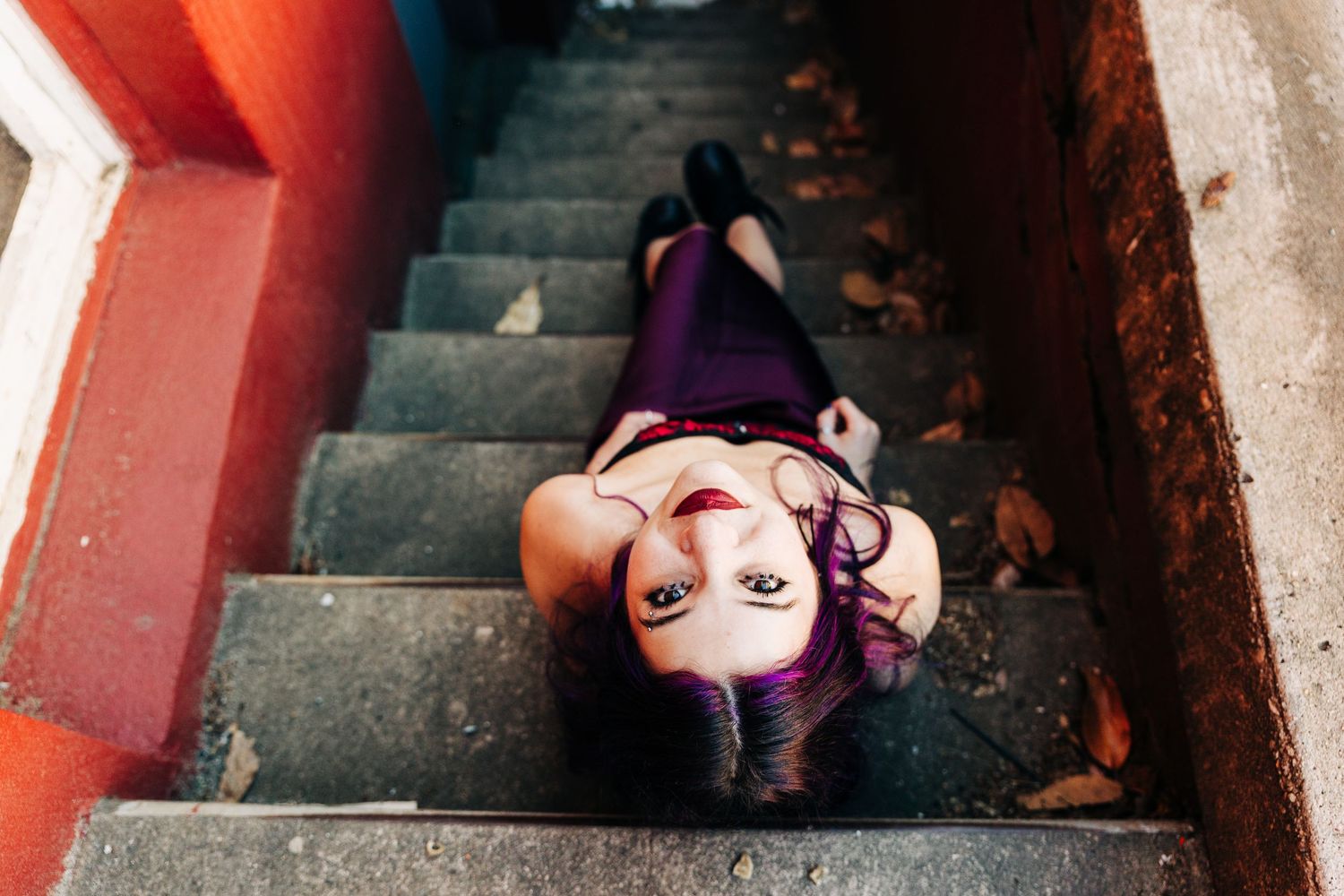 A person wearing purple clothing lies on concrete stairs surrounded by autumn leaves and red painted walls.
