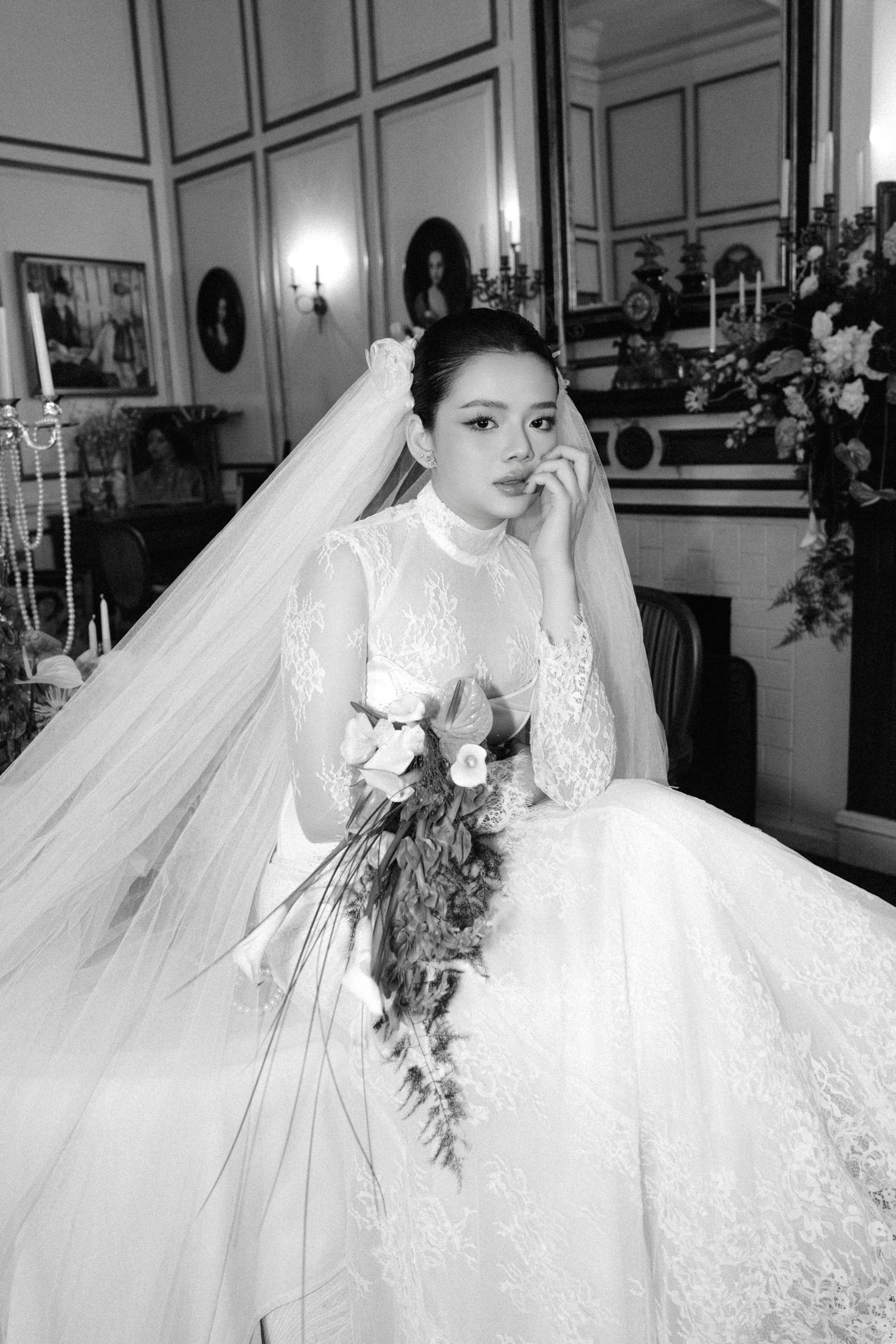 Black and white portrait of a bride in a ball gown with veil and bouquet in an ornate room.