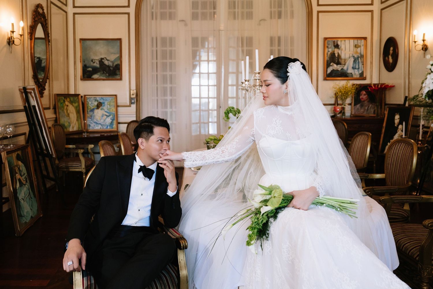 Bride and groom share an intimate moment, she touches his face while holding a green bouquet in an elegant room.