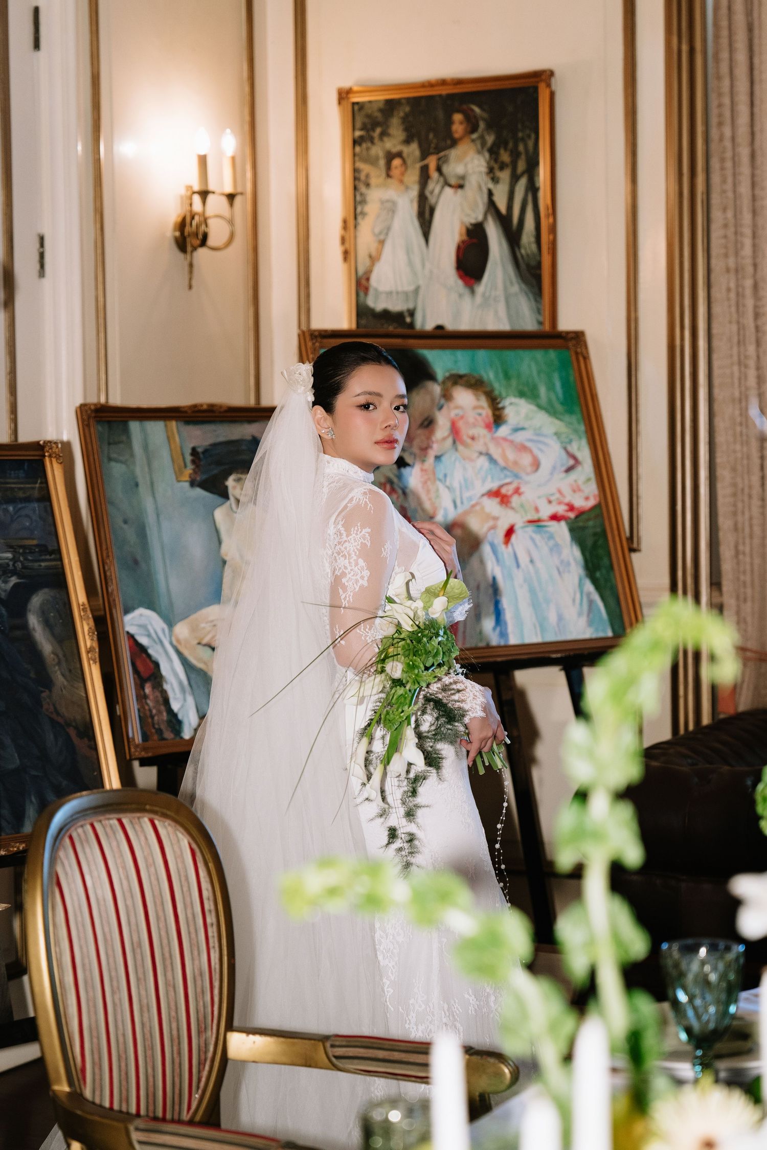 Elegant bride in lace gown holds a green bouquet while standing among classic oil paintings in a vintage room.