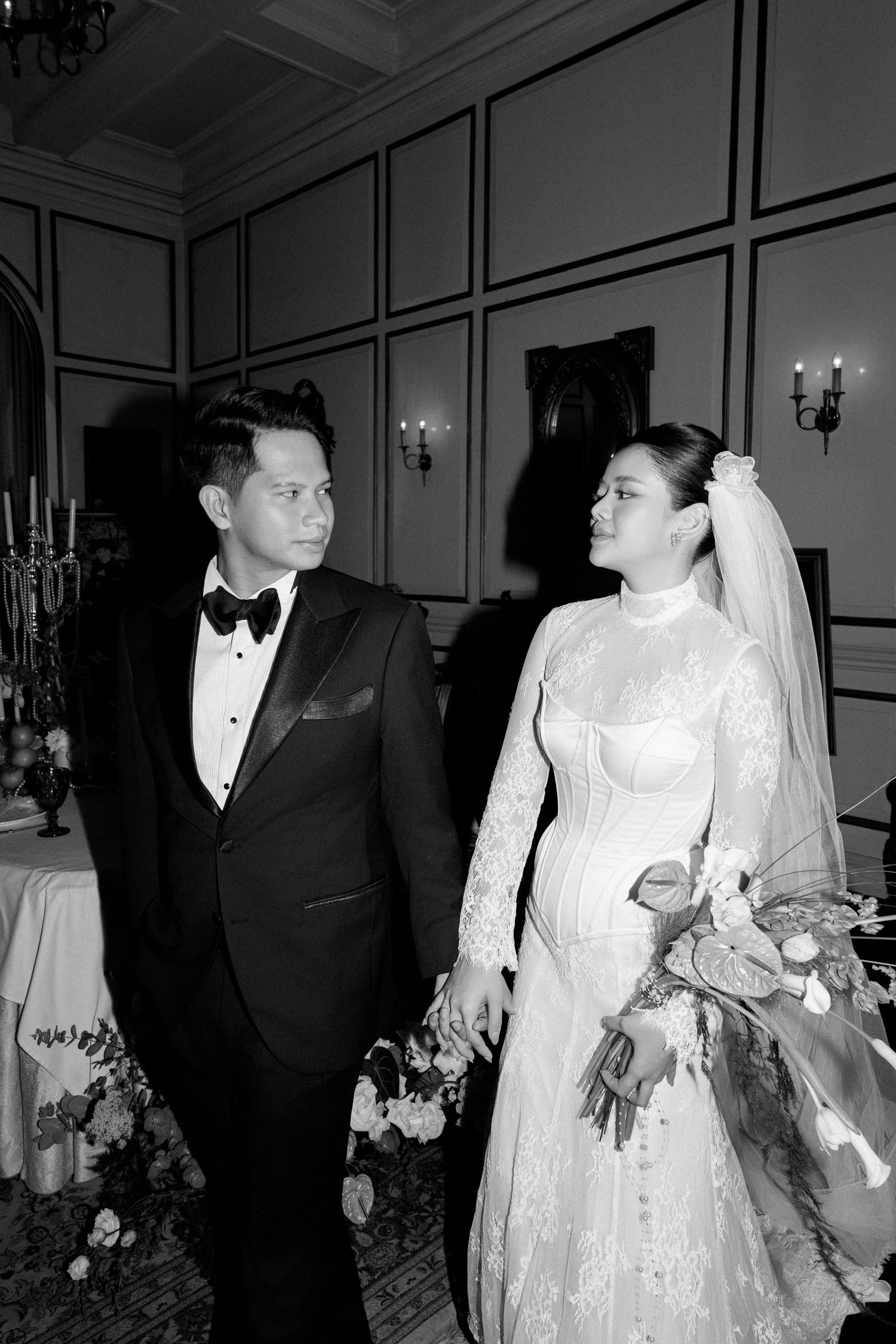 Black and white portrait of bride and groom in formal attire holding hands and gazing at each other.