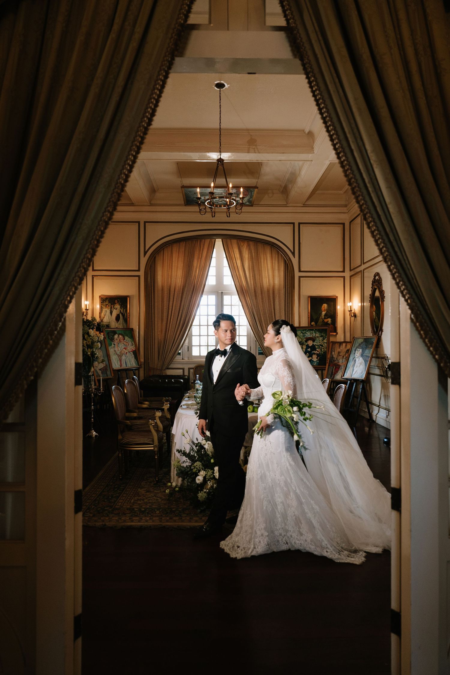 Bride and groom hold hands in a grand vintage hall framed by dramatic drapes and a chandelier overhead.