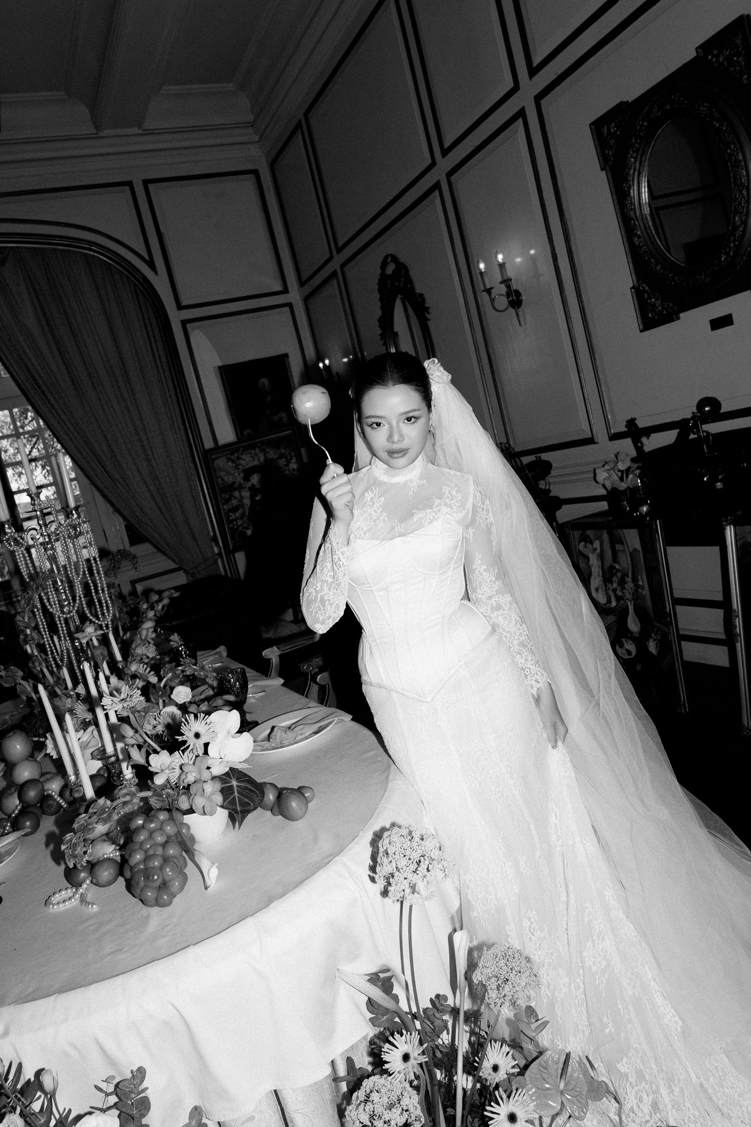 Black and white photo of bride in lace gown holding a decorative wand beside an elegant floral reception table.