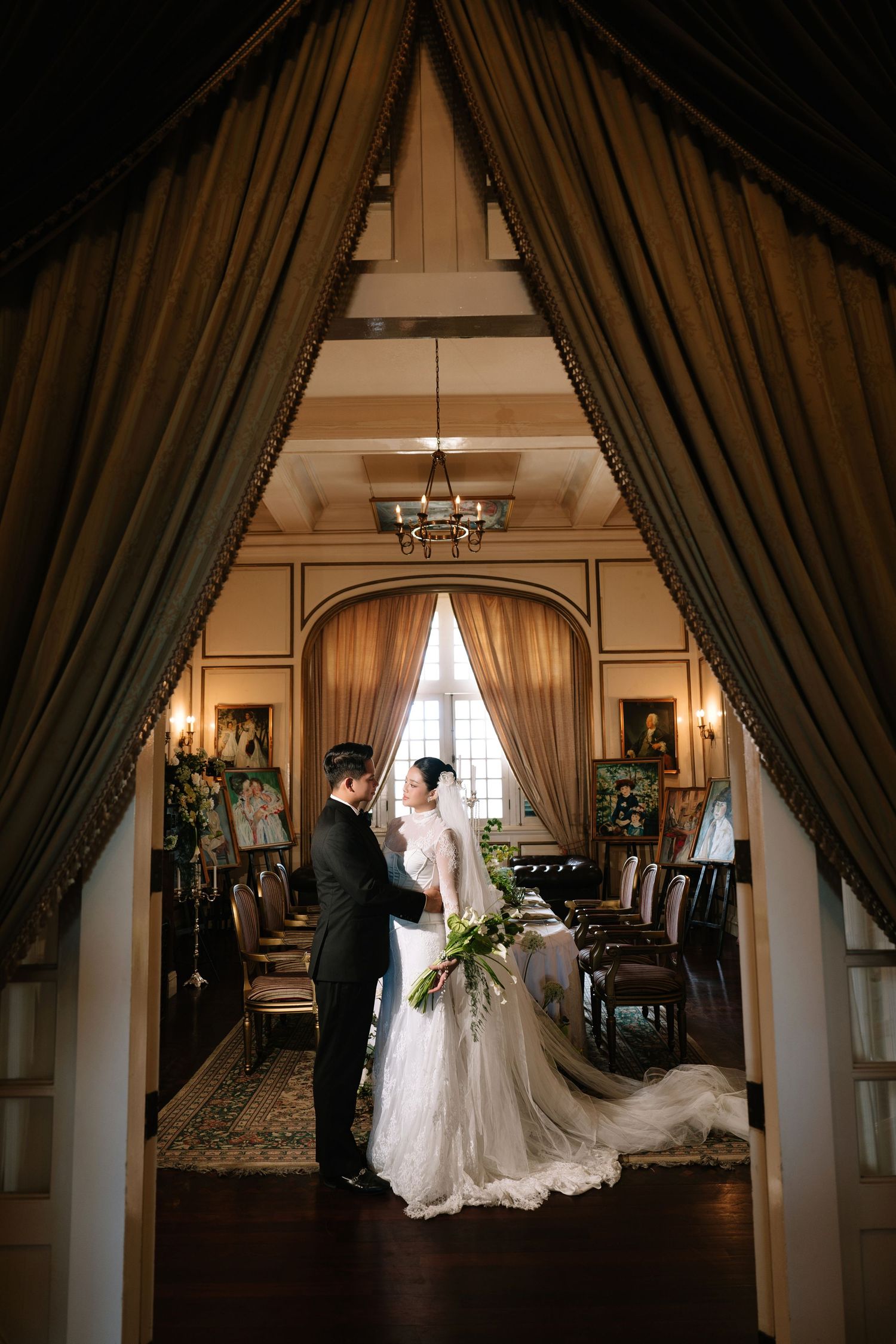 Bride and groom stand facing each other in a grand room framed by floor-length velvet drapes and chandelier.