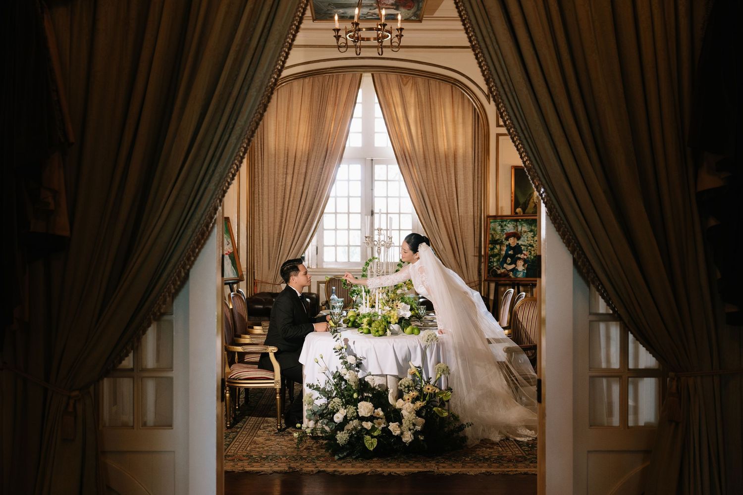 Bride and groom exchange vows at a floral-adorned table framed by elegant drapes in a classic venue.