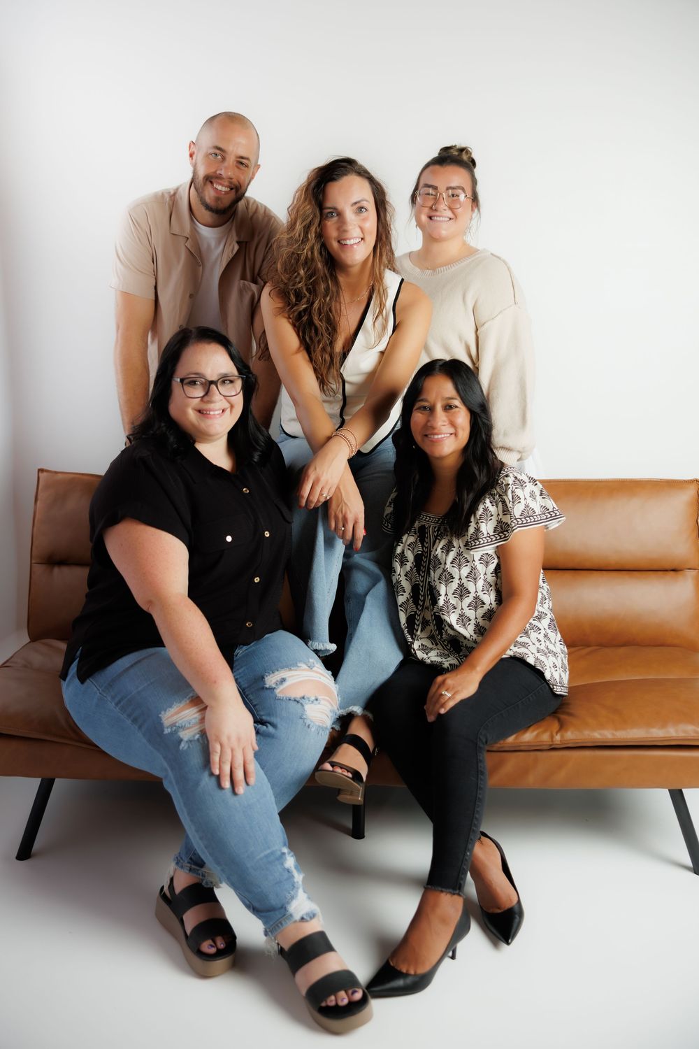 Five friends pose casually on a brown leather couch against a white studio backdrop.