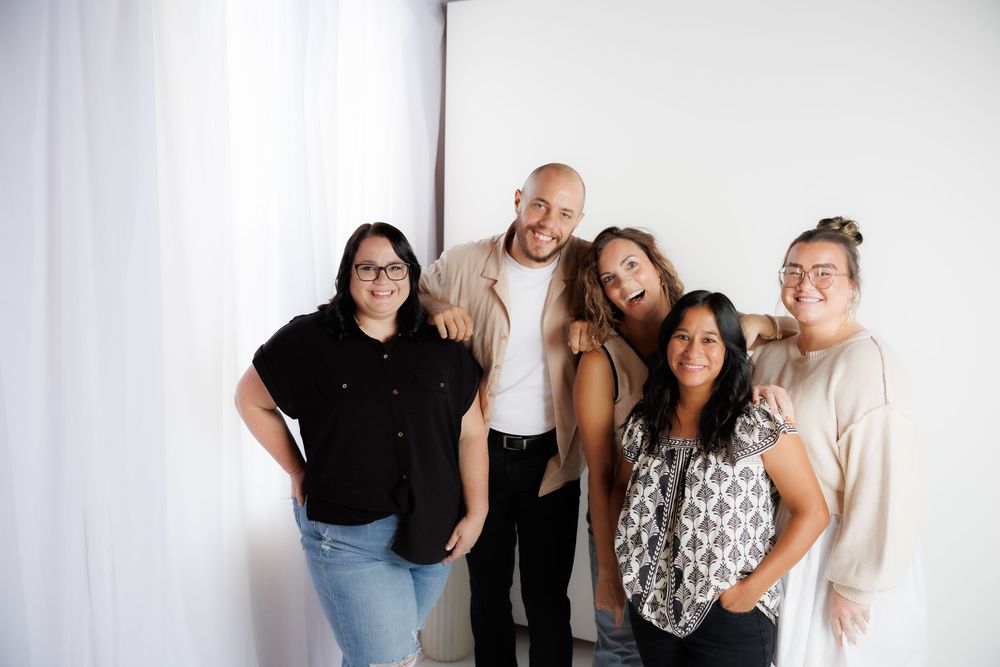A group of five people pose together casually against a white backdrop in a studio setting.