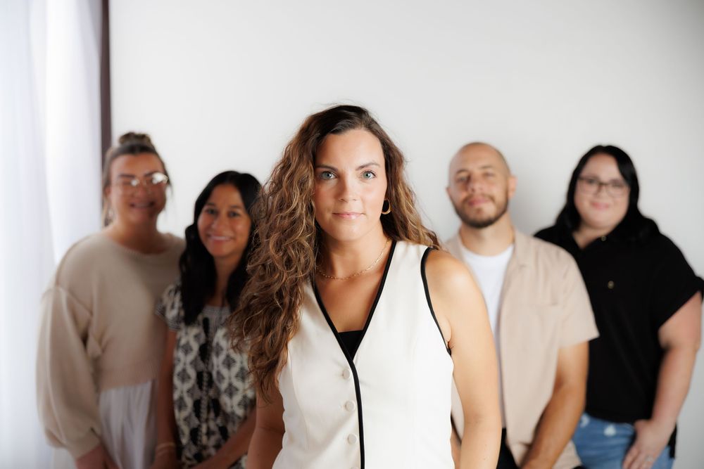 A group of professional people stand together in a row wearing neutral colors against a minimalist white background.