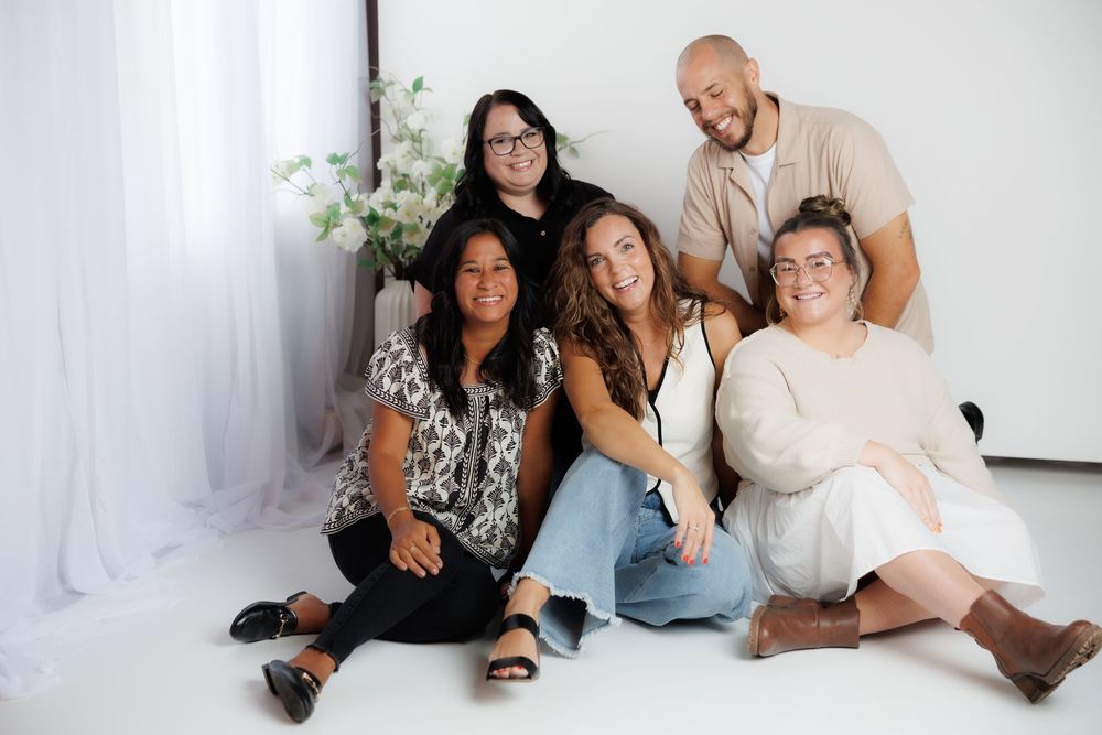 A diverse group of five people sit close together on the floor against a white backdrop with sheer curtains and florals.