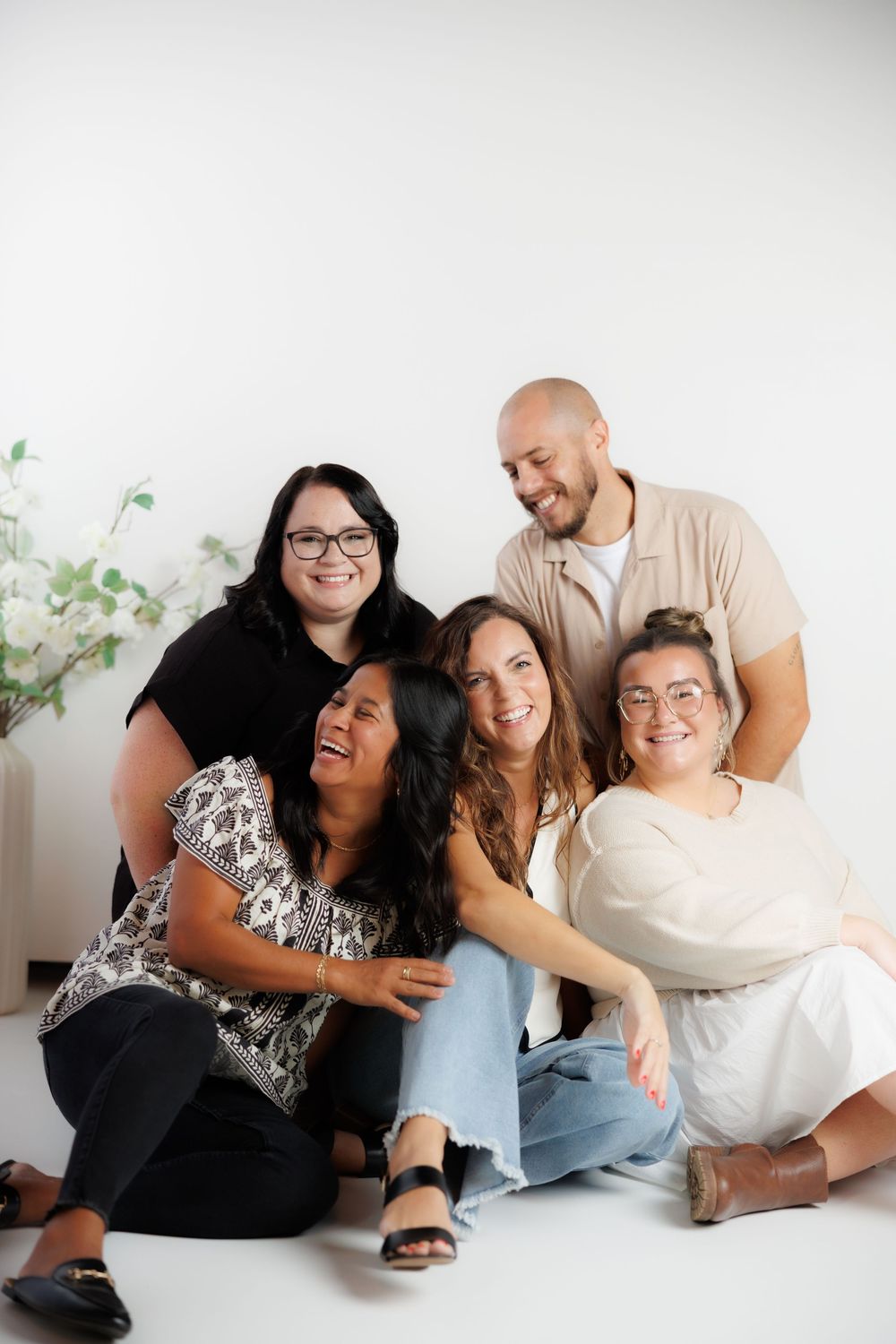 A group of diverse friends laughing and embracing while sitting together against a white wall with green foliage.