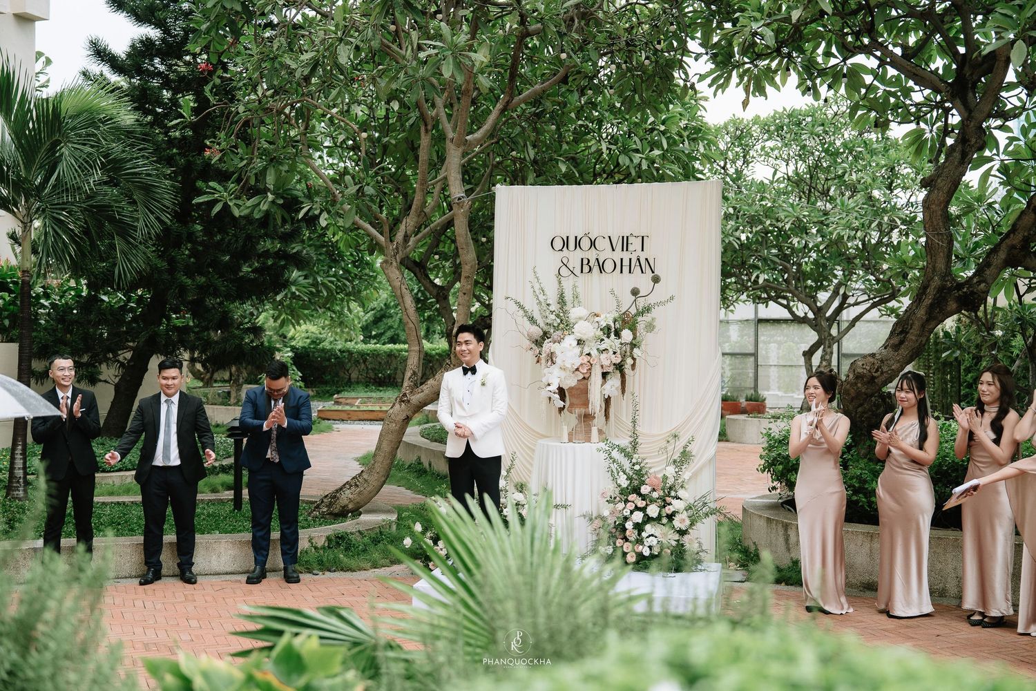 Wedding guests applaud as couple stands before elegant white floral backdrop at outdoor garden ceremony.