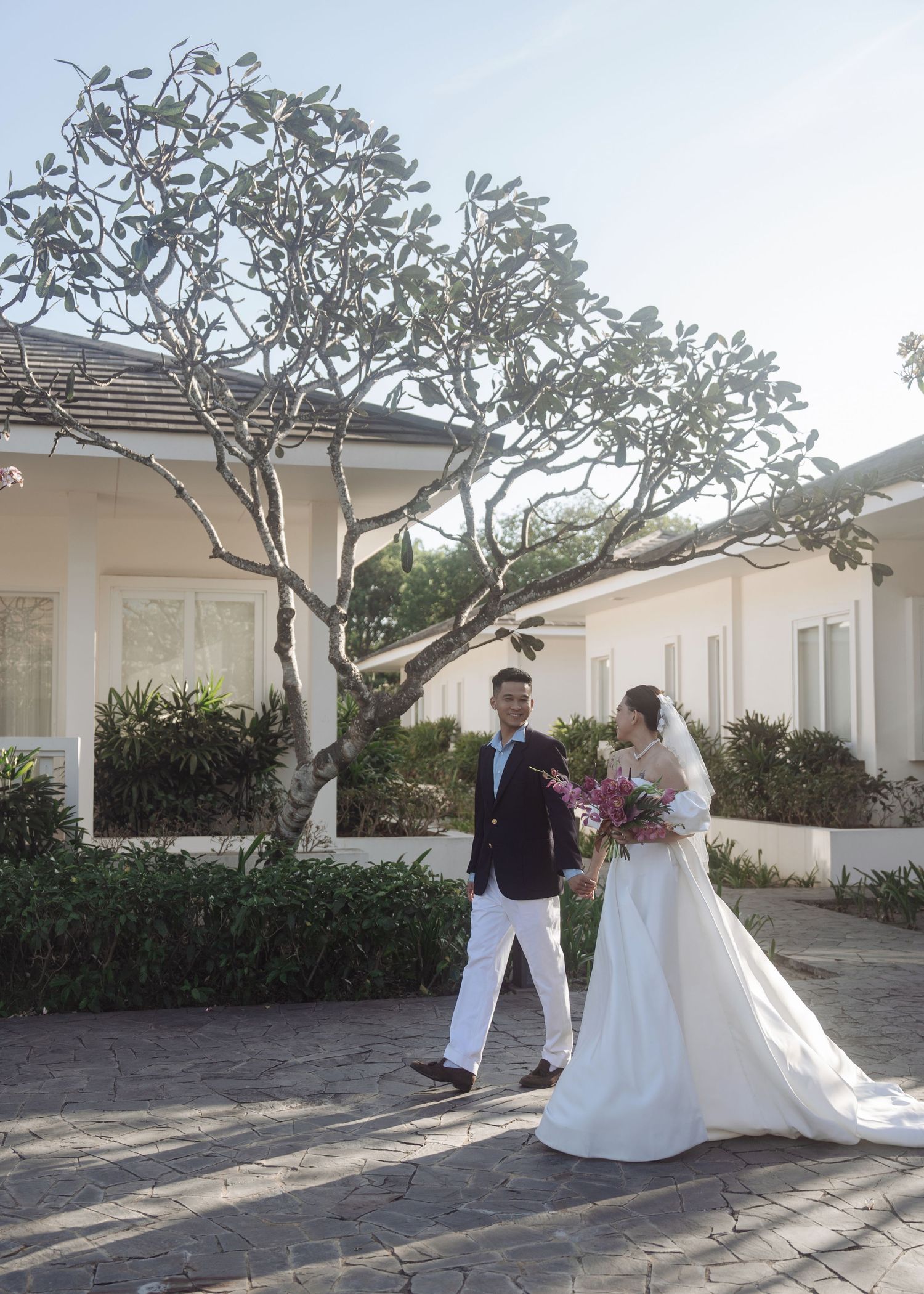 Bride and groom in formal wedding attire walk hand in hand outside white villa surrounded by trees.
