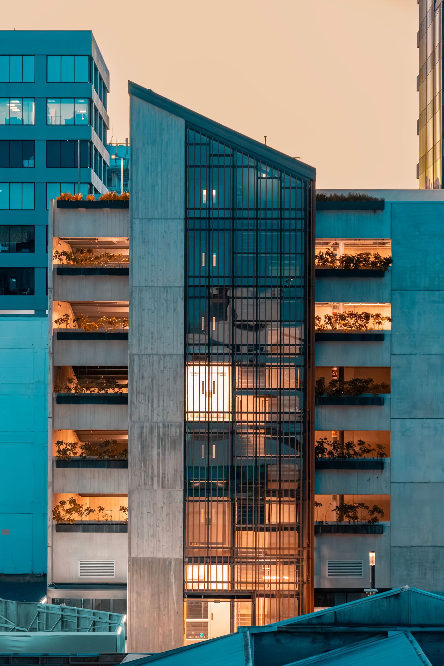 Tall concrete building with a glass centre column and greenery-filled terraces, warmly illuminated at dusk.