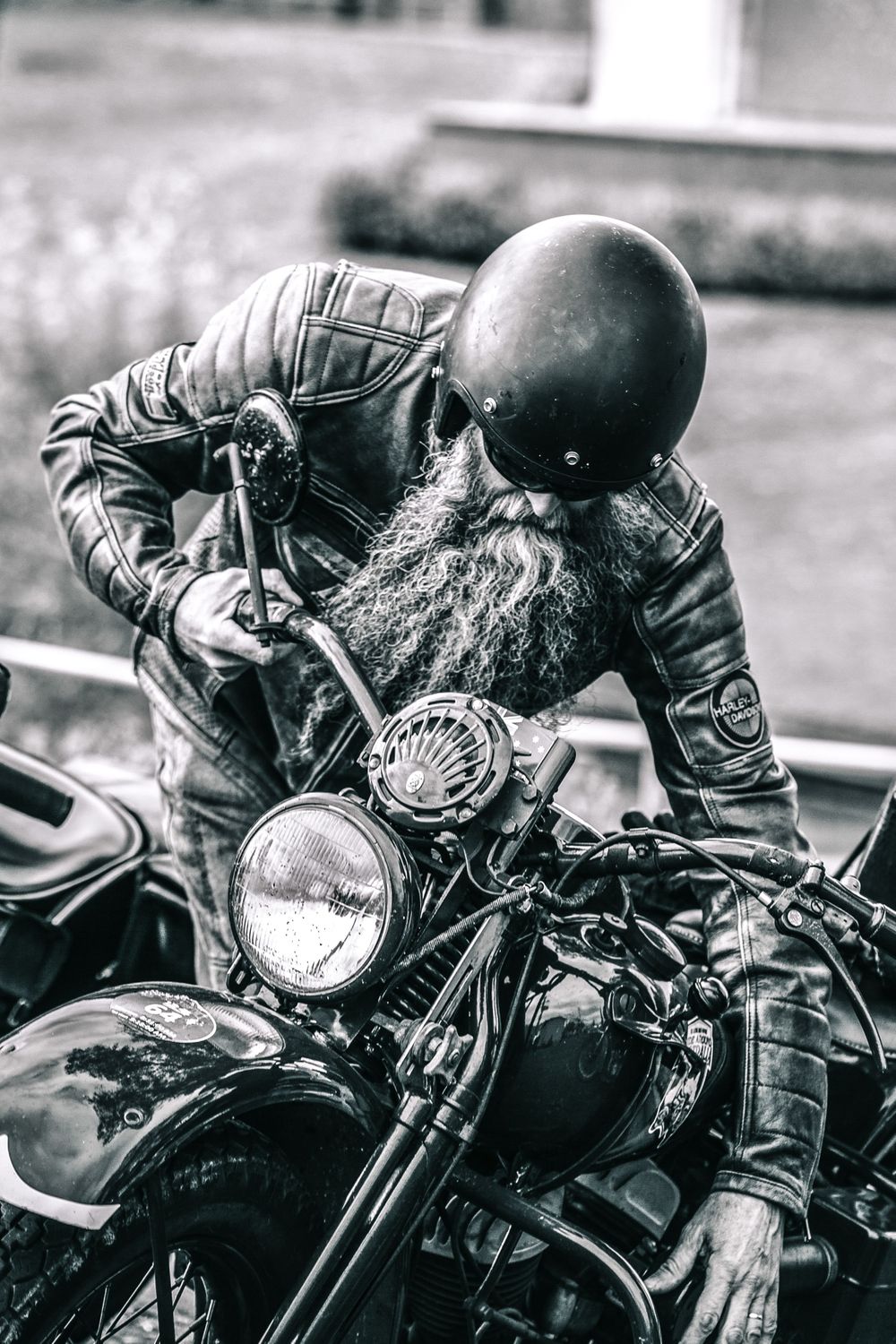 Black and white photo of a bearded biker in leather jacket and helmet leaning over a vintage motorcycle.