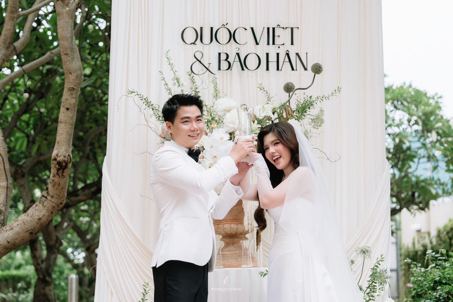 Smiling couple in white attire toast champagne before Quoc Viet and Bao Han wedding backdrop.