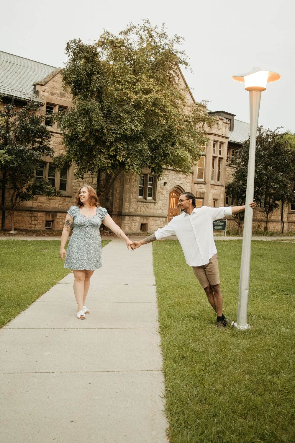 Couple holding hands on campus walkway, man leaning on lamppost, woman in floral dress laughing.