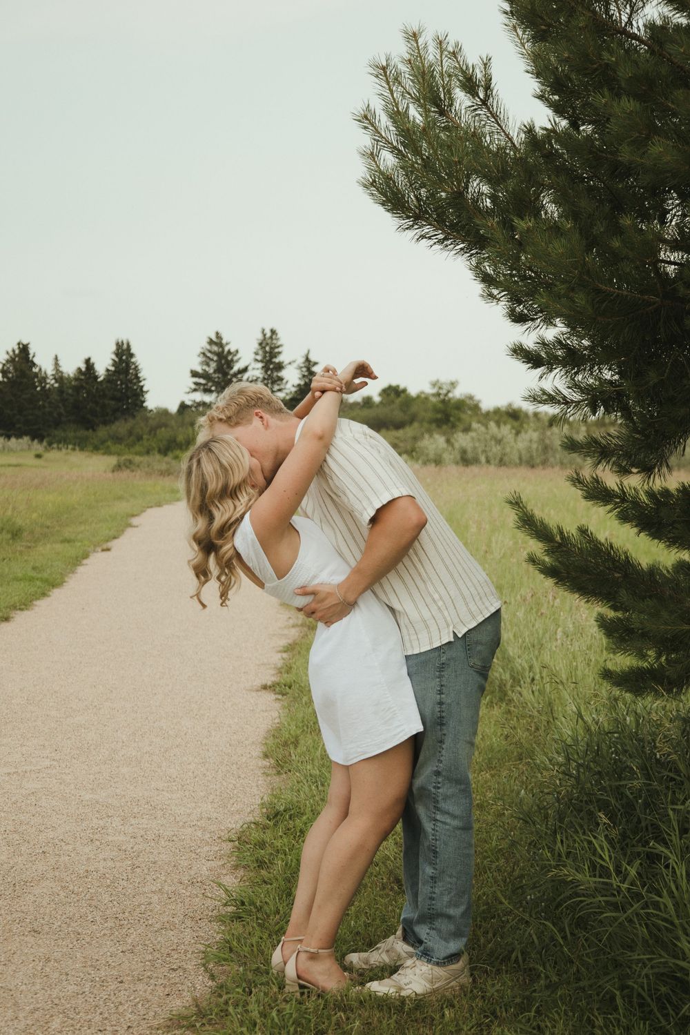 Couple embracing and kissing on a scenic path surrounded by green trees and grass on a warm summer day.