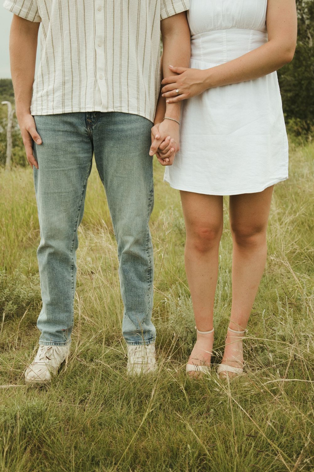 Couple holding hands in a grassy field, wearing casual summer outfits including jeans and a white dress.