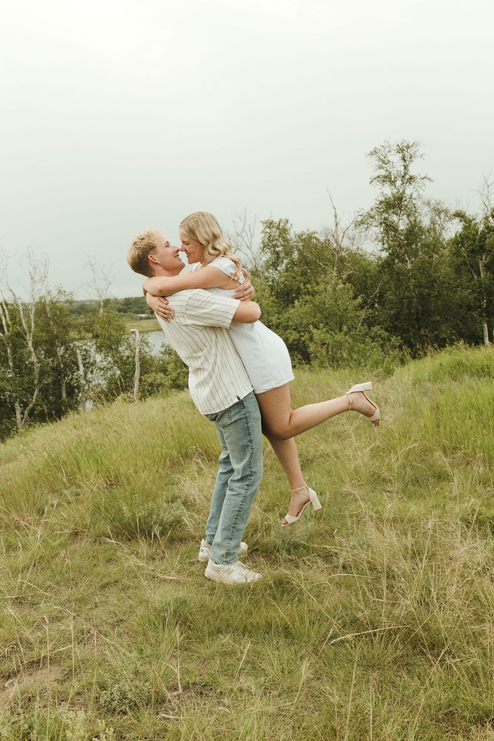 A couple embraces and kisses outdoors on a grassy hill, the man lifting the woman in a white dress.