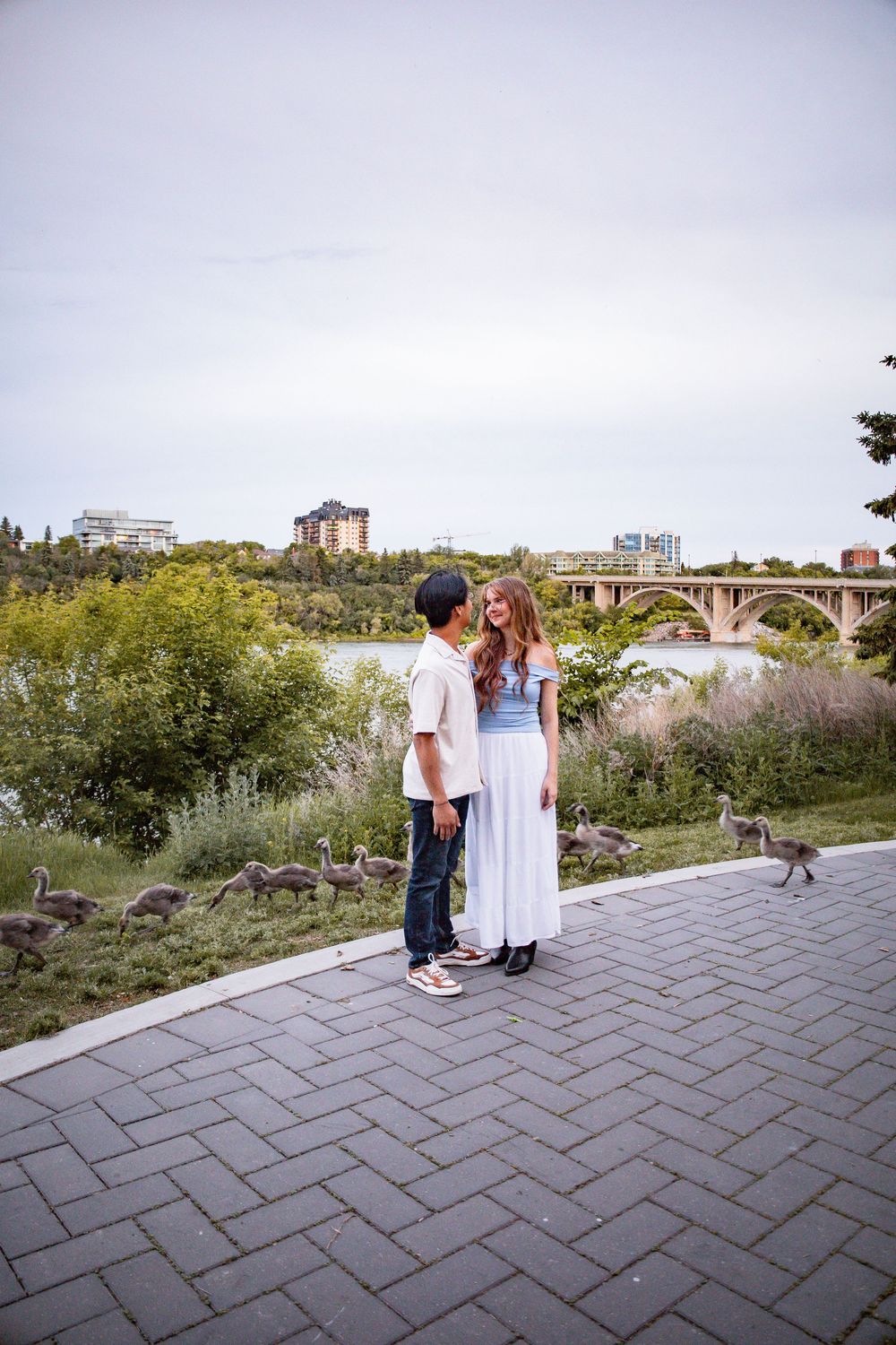 Couple standing on a brick path by a river with geese and a bridge backdrop.