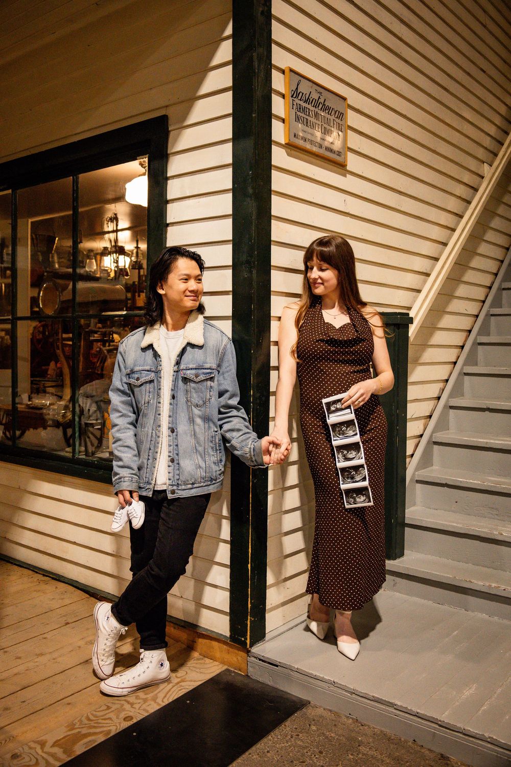 Couple holding hands outside a vintage shop, woman in polka dot dress holding ultrasound photos, announcing pregnancy.