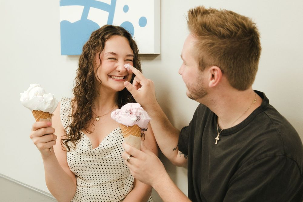 A smiling couple sharing ice cream cones, with a man playfully touching a woman's nose with a pink scoop.