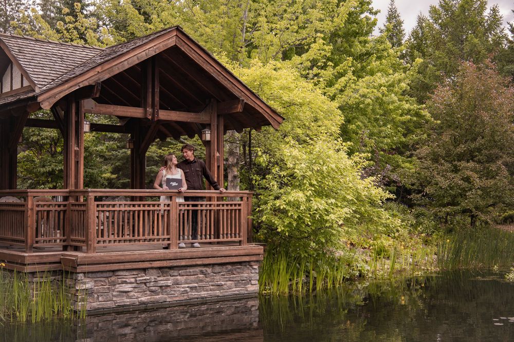 Couple standing on a wooden gazebo overlooking a serene pond surrounded by lush green trees in a garden.