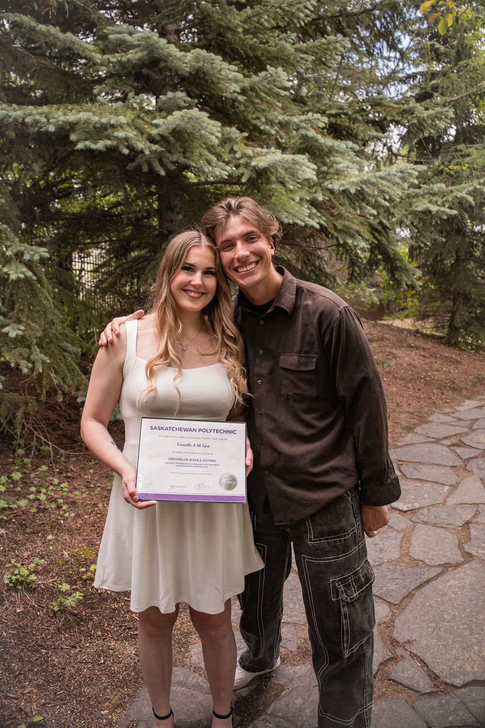 Young woman in white dress holds Saskatchewan Polytechnic certificate, posing with a man outdoors near evergreen trees.