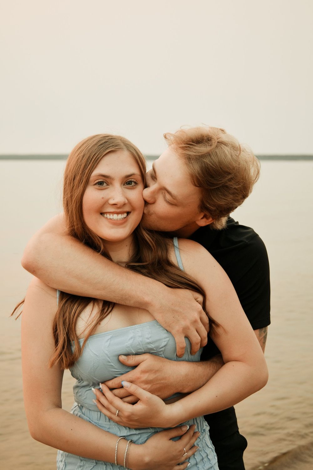 A young couple sharing a tender moment, man kissing woman's cheek while embracing near a lake.