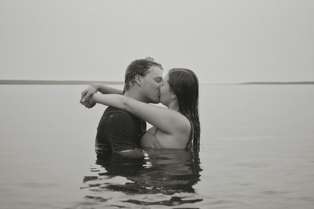 Couple sharing a romantic kiss while standing in calm open water in a black and white outdoor photo.