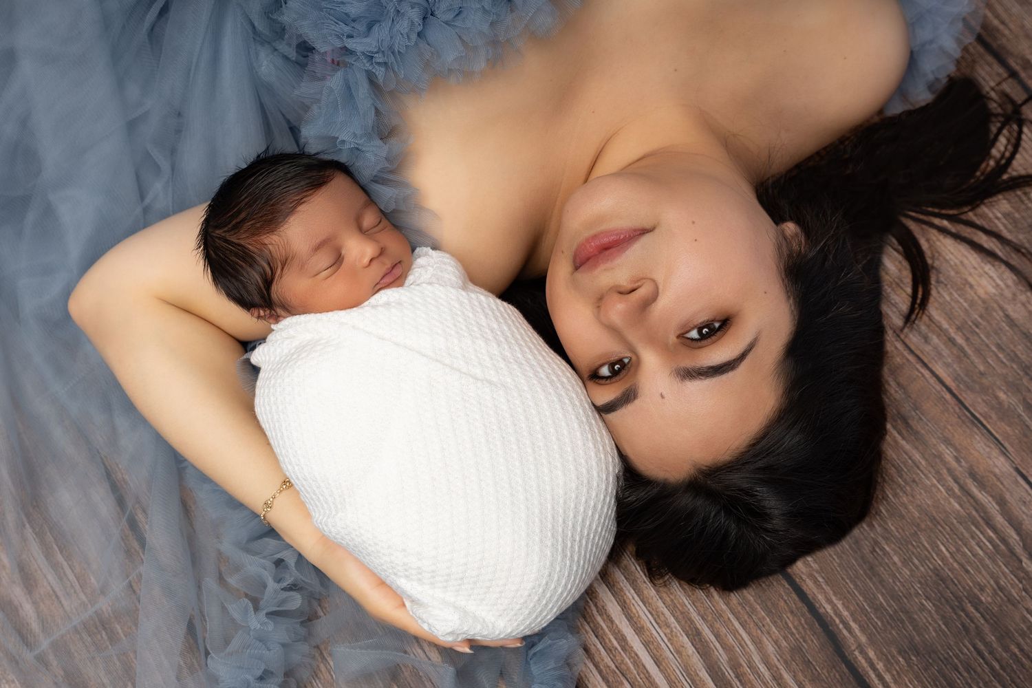 A young couple in casual attire lovingly holds their swaddled newborn baby against a gray studio background.