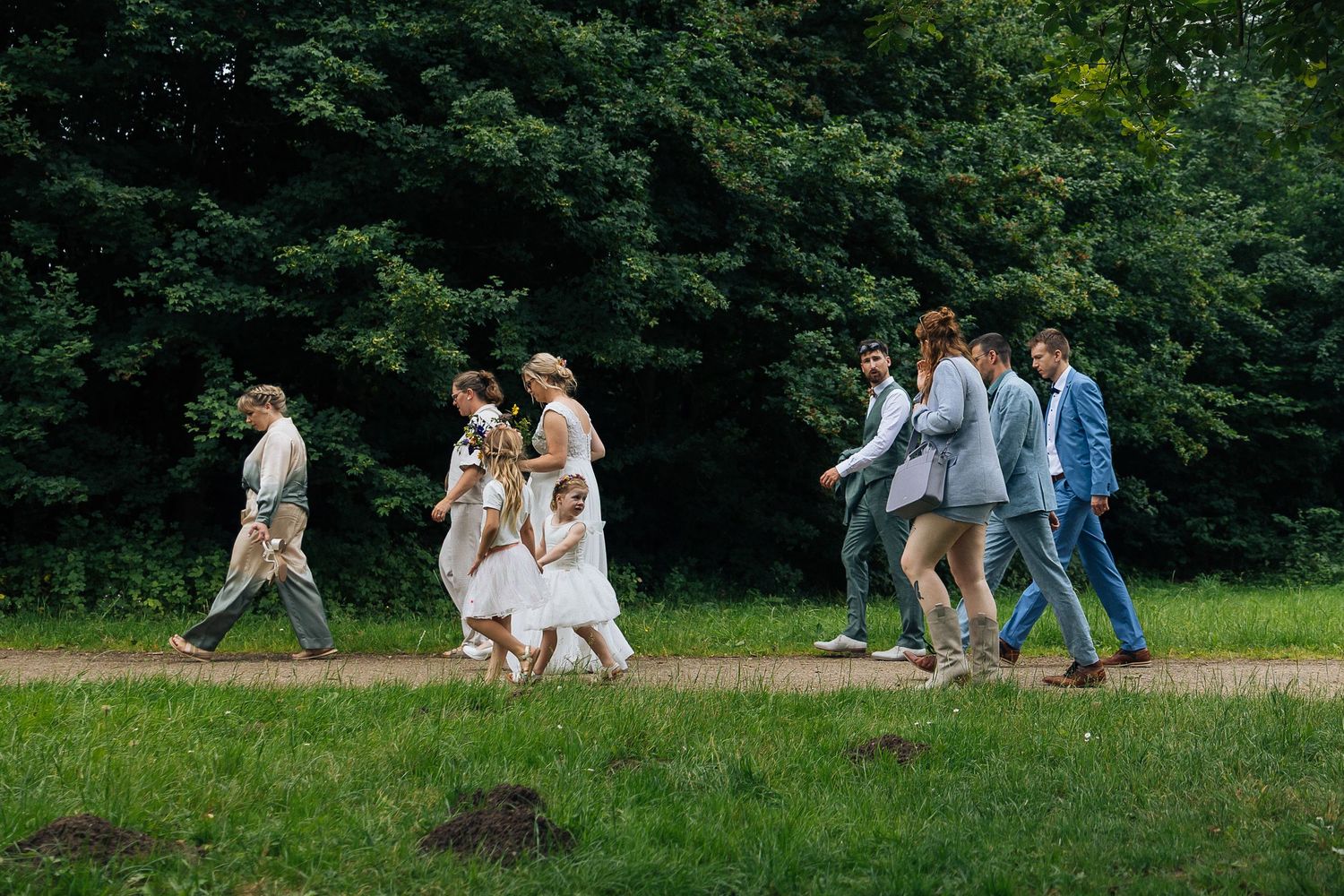 Wedding party walks together across a green field surrounded by trees.
