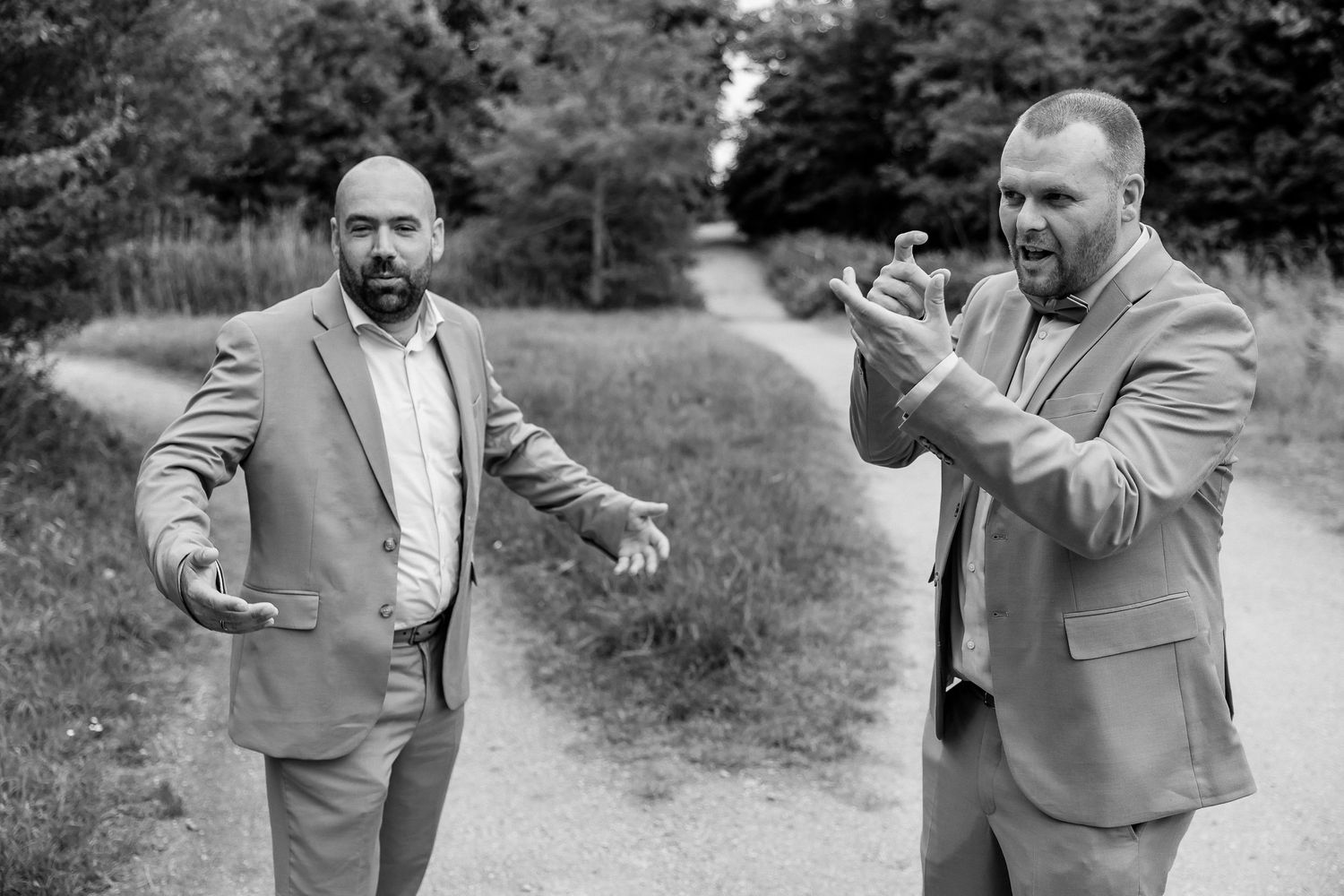 Two men in light gray suits gesturing and laughing on a nature path.