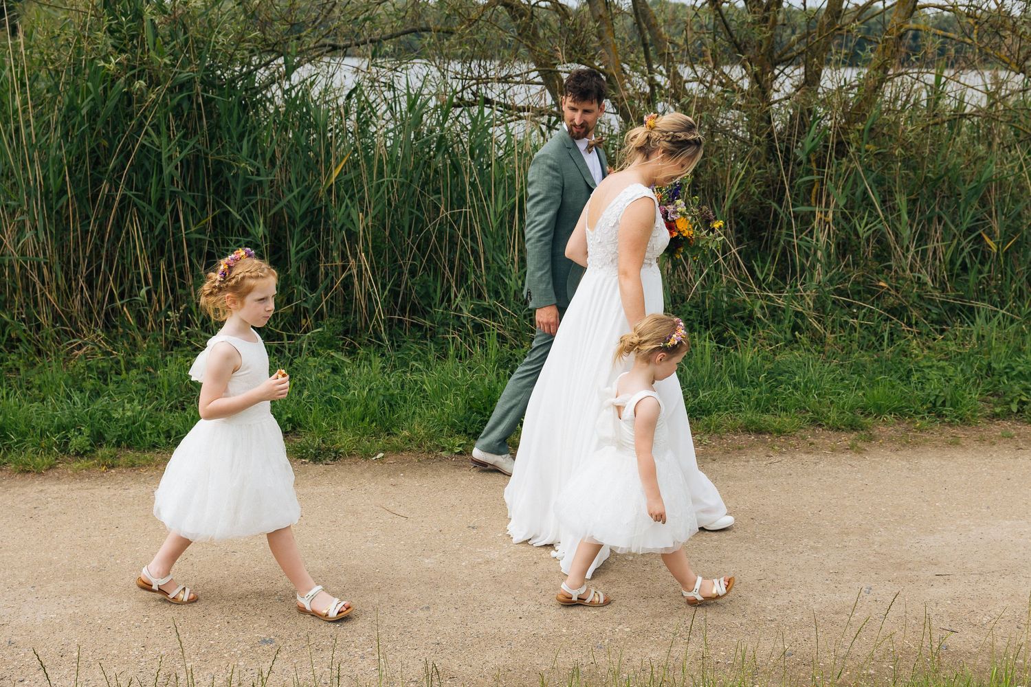 Family walking on sandy path beside tall reeds near water.