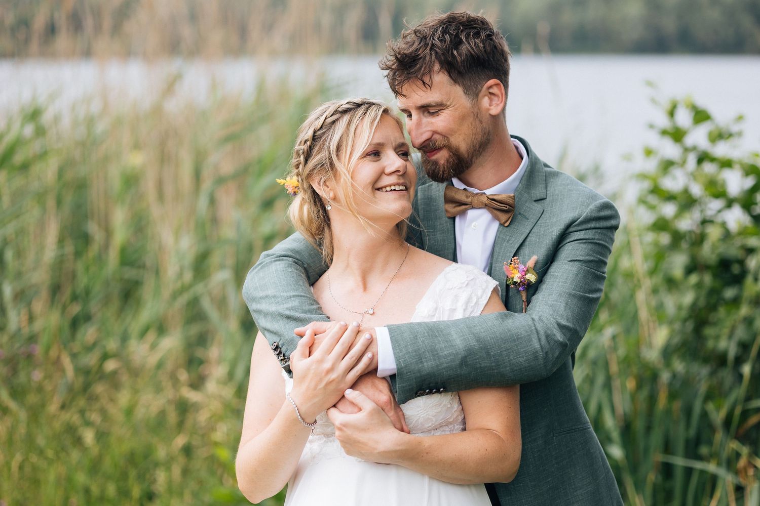 Couple embracing in romantic moment by lake with tall grasses.