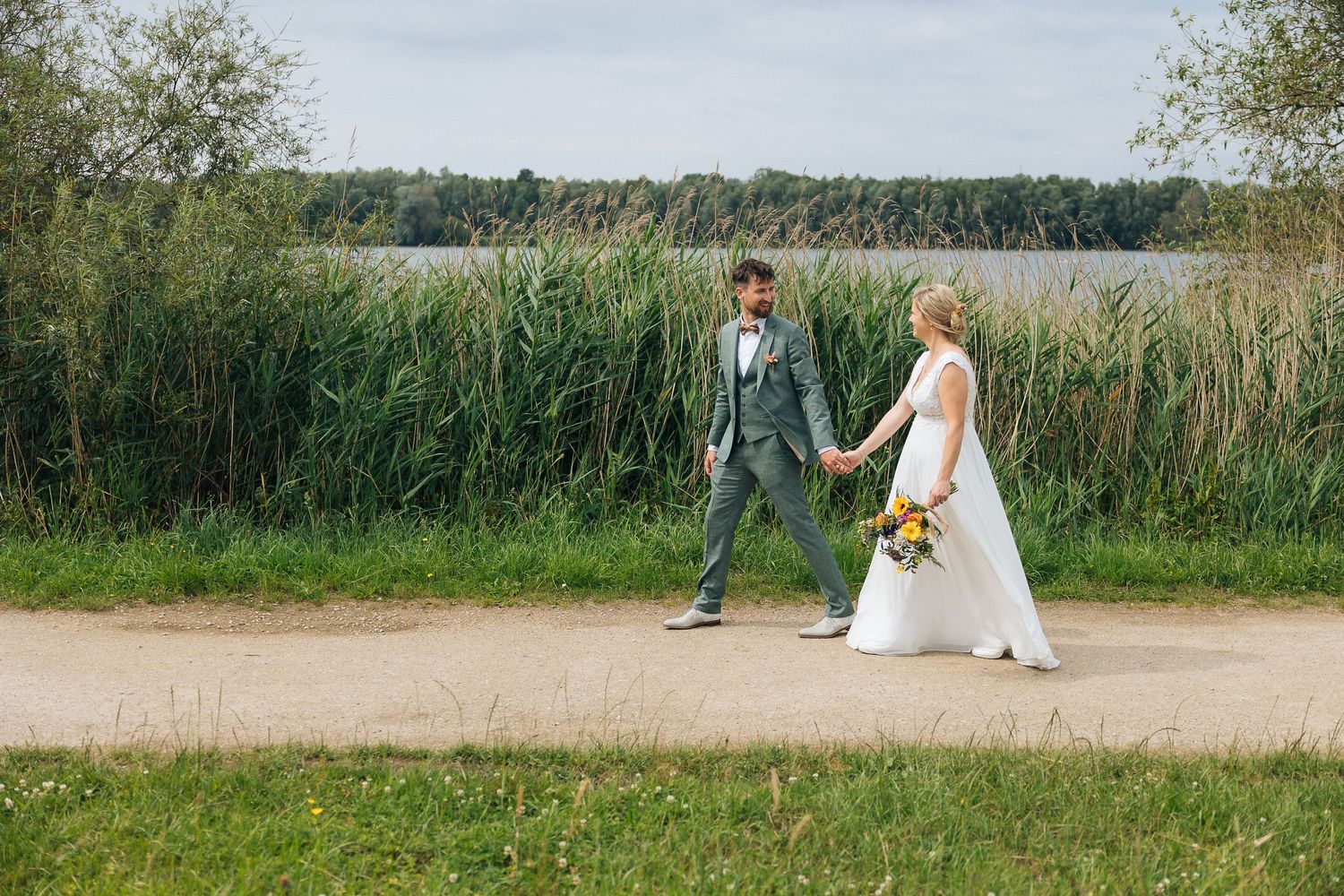Couple walking on path beside tall reeds during outdoor wedding shoot.