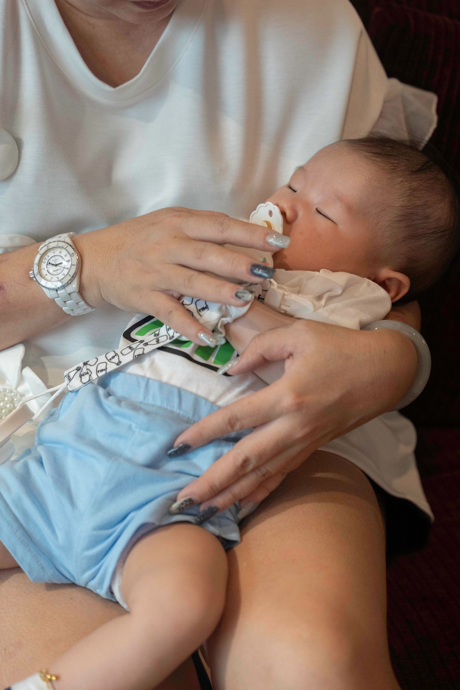 A baby in light blue clothing drinks from a bottle while being held in someone's lap wearing a white shirt and watch.