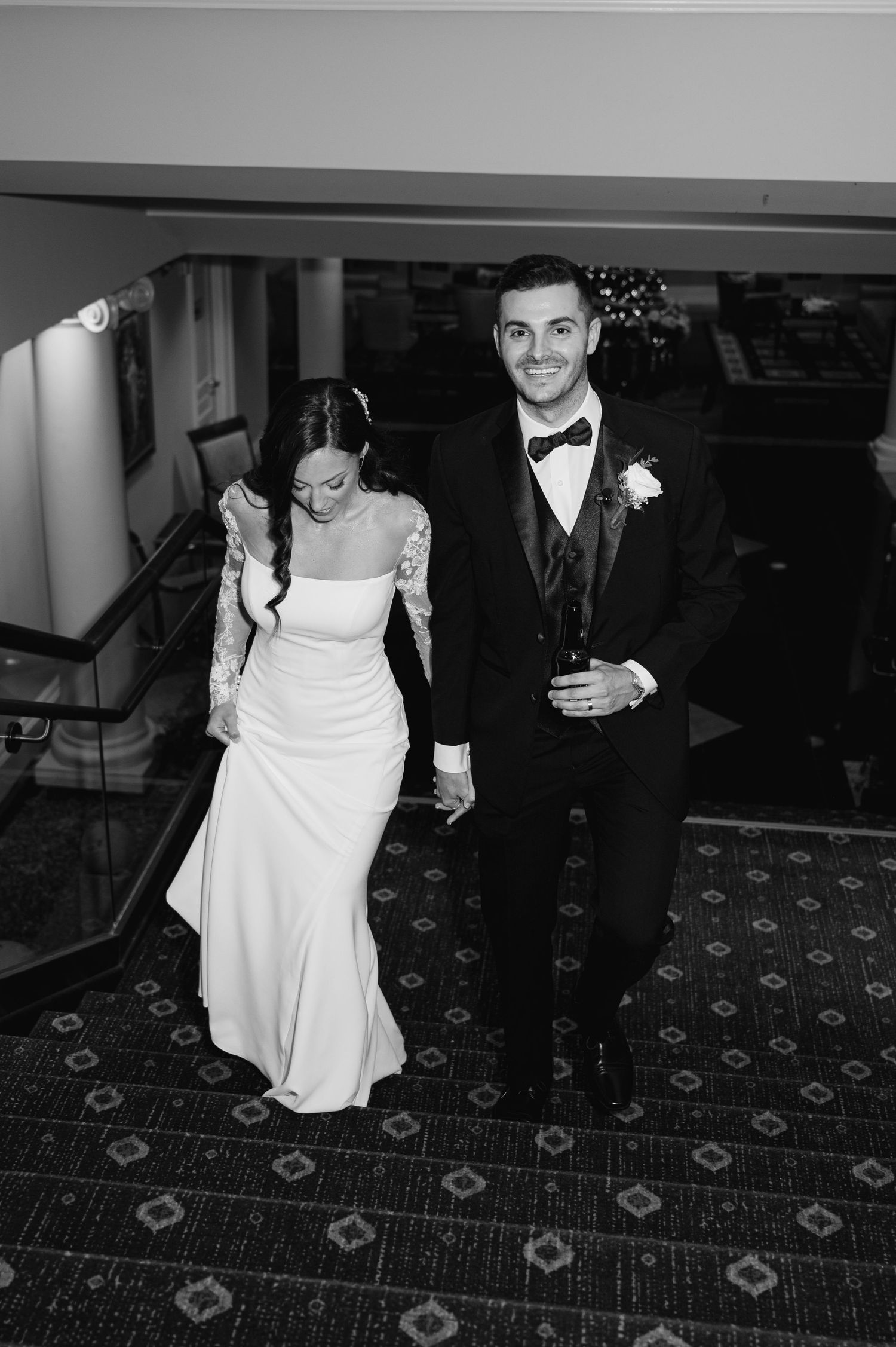 A black and white wedding photo of a bride in white dress and groom in tuxedo walking up  carpeted stairs towards their cocktail hour.