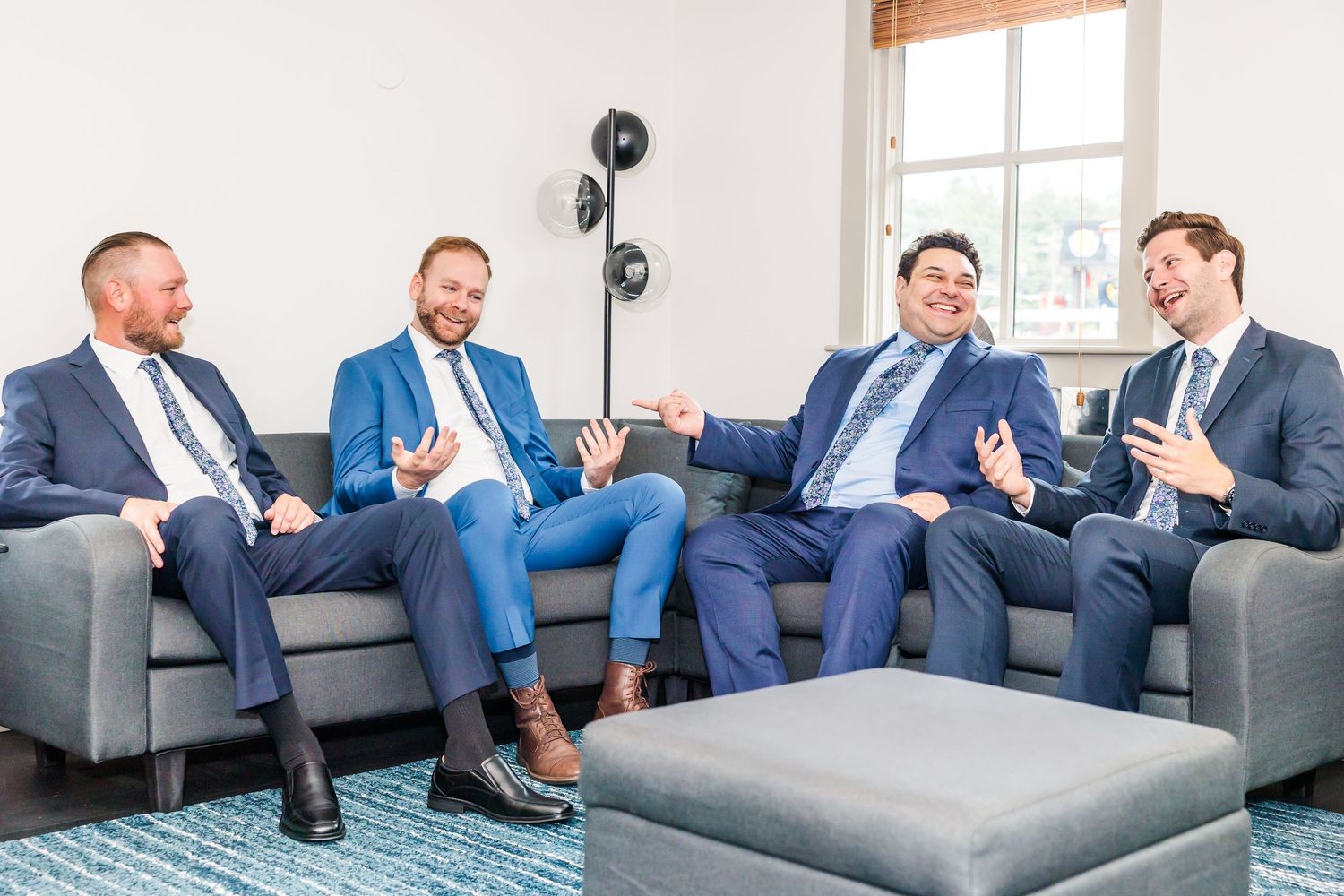 Four men in business suits relax and laugh while sitting on a gray couch in a modern office setting.