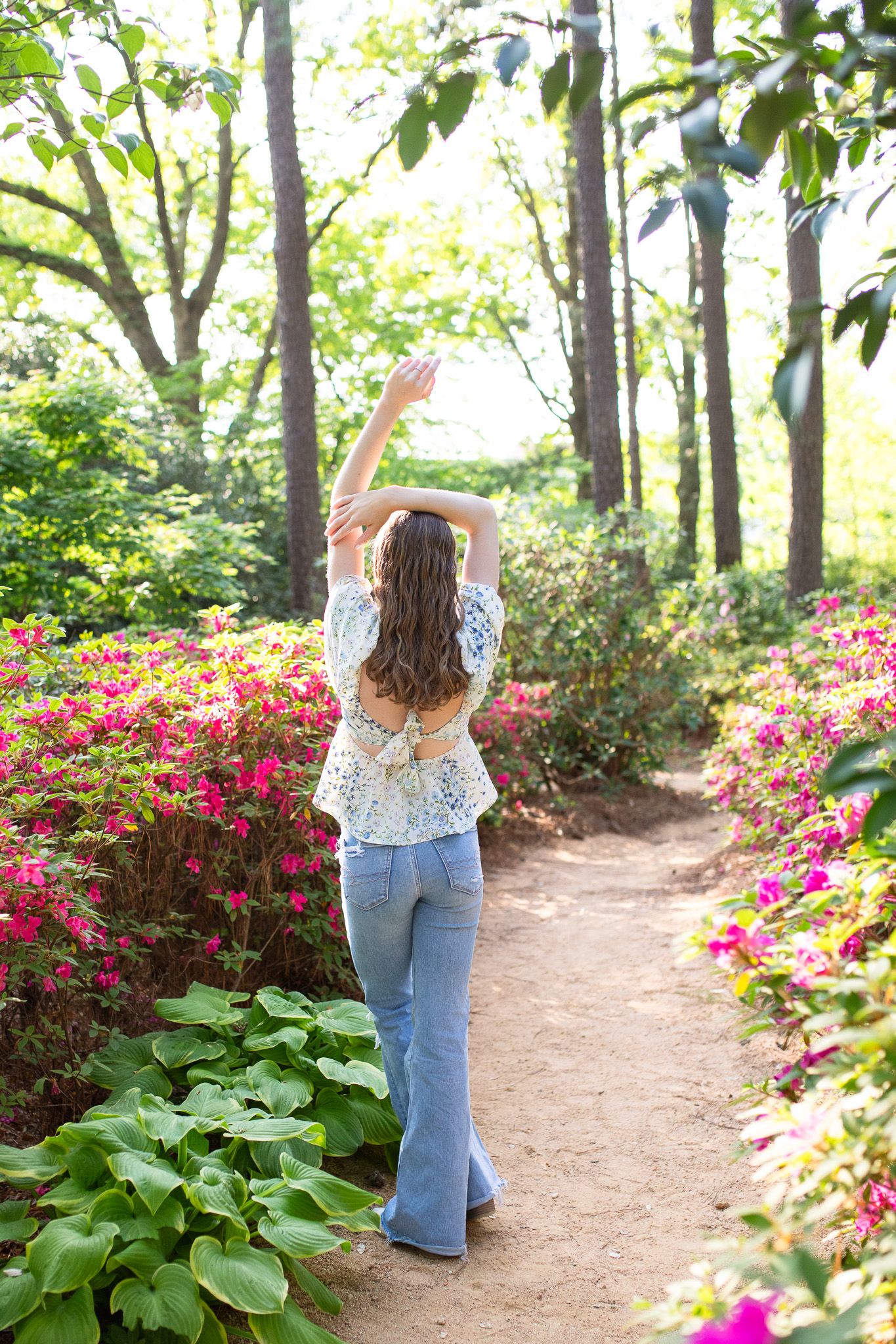 A senior grad walks along a garden path surrounded by vibrant pink azaleas and lush greenery on a sunny spring day.