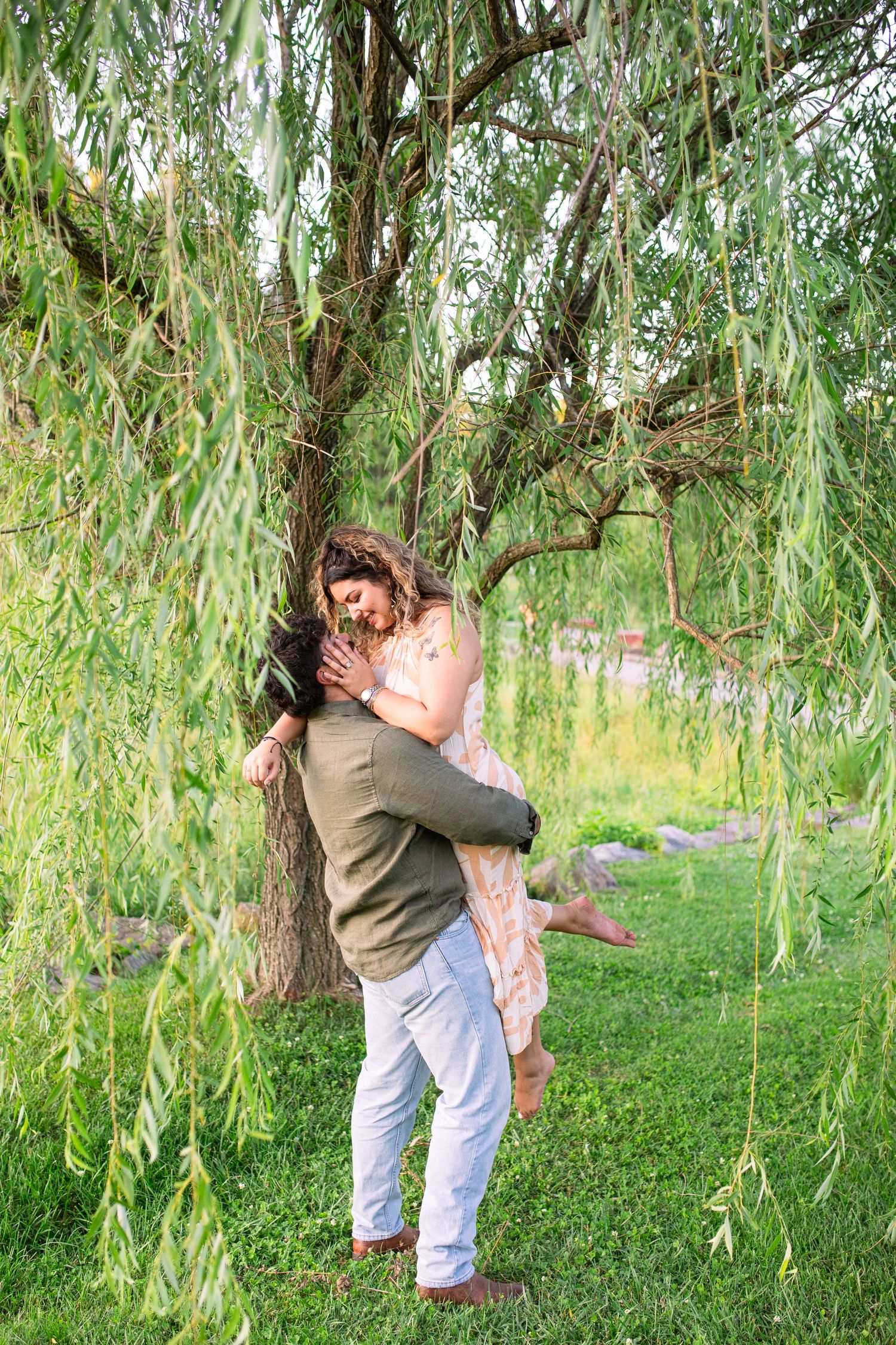 A romantic couple embraces beneath drooping willow branches in a lush summer garden setting at NCMA